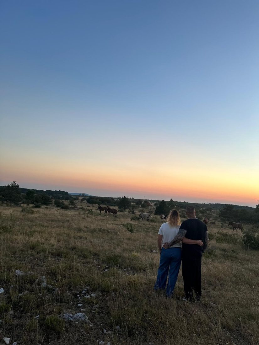 A couple standing close together watching a herd of horses at sunset